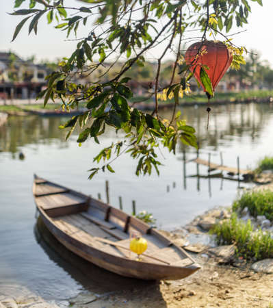 Scenic view of green tree branches decorated with traditional red lantern in Hoi An Ancient Town (Hoian), Vietnam. Wooden boat parked on bank of the Thu Bon River. Focus on the foliage.の写真素材
