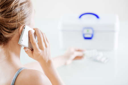 Closeup view of woman calling to doctor for getting medical consultation on phone at home. White plastic box with first aid kit and pills in blister packs on table.の写真素材