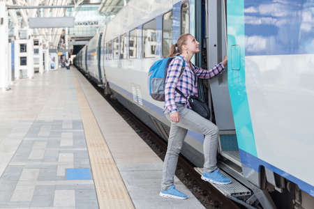 Young female tourist entering train. Smiling pretty woman with blue backpack at railway station....