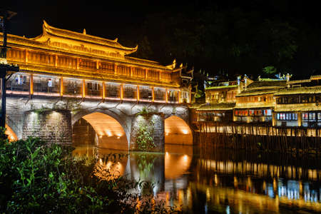 Scenic night view of the Hong Bridge (Rainbow Bridge) over the Tuojiang River in Phoenix Ancient Town (Fenghuang), China. Old traditional Chinese wooden riverside houses on stilts reflected in water.の写真素材