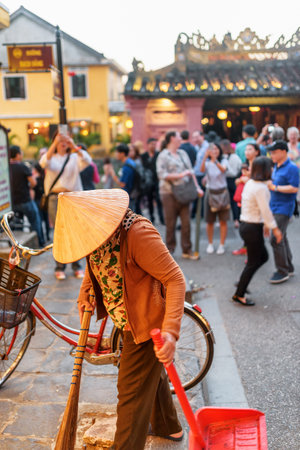 Hoi An (Hoian), Vietnam - April 11, 2018: Vietnamese janitor in traditional bamboo hat working on street of Hoi An Ancient Town. Tourists taking pictures on the Japanese Covered Bridge background.のeditorial素材
