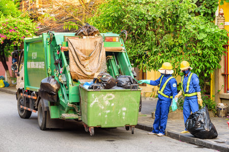 Hoi An (Hoian), Vietnam - April 12, 2018: Two garbagemen loading trash by hand. Garbage truck at old street of Hoi An Ancient Town in morning. Hoian is a popular tourist destination of Asia.のeditorial素材
