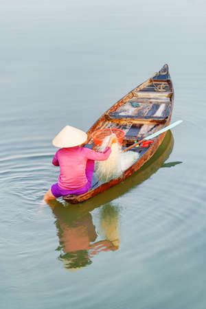 Awesome view of Vietnamese woman in traditional hat on wooden boat checking her fishing net on the Thu Bon River at Hoi An Ancient Town, Vietnam. Hoian is a popular tourist destination of Asia.の写真素材