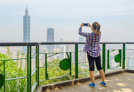 Young female tourist taking pictures and enjoying scenic view of Taipei from top of mountain. Awesome Taipei skyline, Taiwan. Wonderful cityscape. Skyscrapers and other modern buildings of downtown.の写真素材