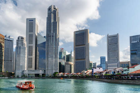 Traditional tourist boat sailing along Boat Quay with azure water in Singapore. Amazing modern buildings are visible on blue sky background. Scenic summer cityscape.の写真素材