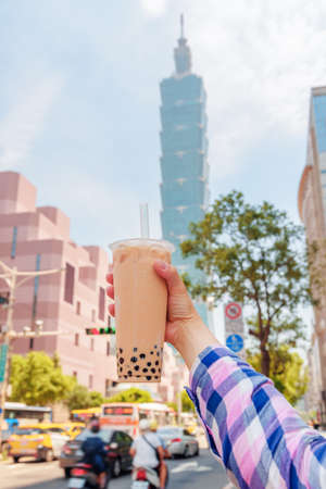 Female hand holding traditional Taiwanese bubble milk tea in downtown of Taipei, Taiwan. Skyscraper is visible in background. The pearl tea is a popular cold drink with chewy tapioca balls.の写真素材