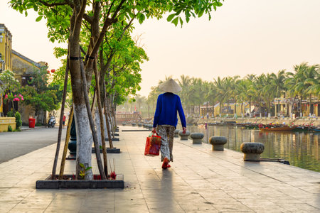 Unknown Vietnamese woman in traditional bamboo hat walking along the Thu Bon River in Hoi An Ancient Town (Hoian) at sunrise, Vietnam. Hoi An is a popular tourist destination of Asia.のeditorial素材