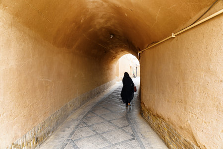 Iranian woman wearing black chador walking along narrow street of the historical city of Yazd. Unique Persian architecture of the ancient town.の写真素材