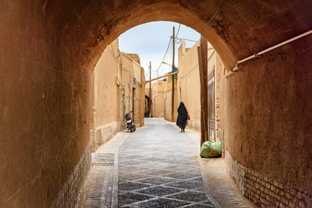 Iranian woman wearing black chador walking along narrow street of the historical city of Yazd. Unique Persian architecture of the ancient town.の写真素材