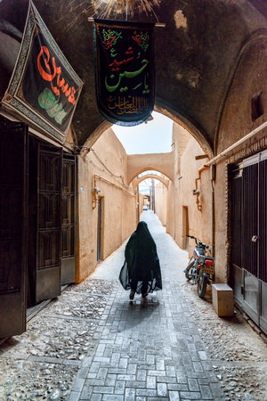 Yazd, Iran - 26 October, 2018: Iranian woman wearing black chador walking along narrow street of the historical city of Yazd. Unique Persian architecture of the ancient town.のeditorial素材