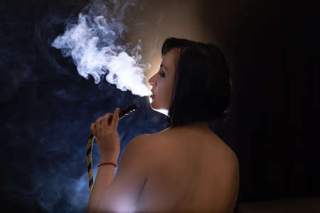 Beautiful, young, naked woman smoking hookah. Attractive girl smoking flavored tobacco. Blow out...