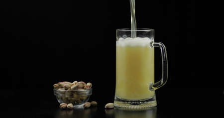 Beer is pouring into glass on black background. Bowl of pistachios nuts.の写真素材
