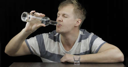 Depressed man drinking vodka from bottle alone in a dark room. Concept of alcoholism. Close-up shotの写真素材