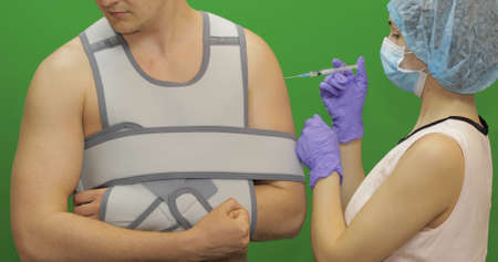 Man with shoulder injury. Nurse with syringe with medicines makes prick from pain. Patient in a bandage for fixing of an elbow joint and a humeral belt. Arm braceの写真素材