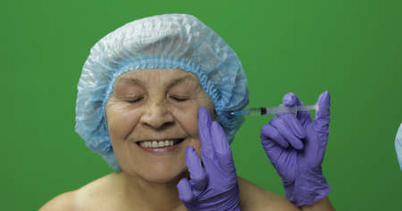 Smiling elderly female in protective hat. Plastic surgeon doctor making facial injections for aging female patient, dermatology. Cosmetic surgery. Green screen. Chroma keyの写真素材
