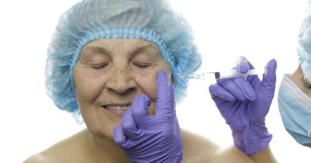 Smiling elderly female in protective hat. Plastic surgeon doctor hands making facial injections for aging female patient, dermatology. Cosmetic surgery. White backgroundの写真素材