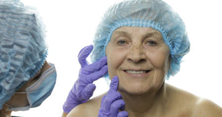 Smiling elderly female in protective hat. Plastic surgeon doctor checking woman face before cosmetic surgery. Dermatology. Doctor advises a patient who agrees with the suggestions. White backgroundの写真素材