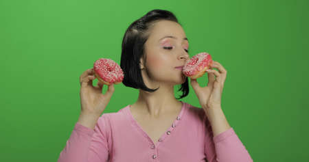 Happy beautiful young girl on a chroma key background having fun with donuts. Cute woman in a pink shirt posing with donuts. Making facesの写真素材