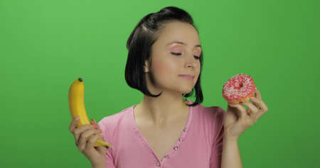 Starting healthy eating. Say no to junk food. Beautiful sad young girl on a chroma key background holds donut in one hand and banana on other. Cute woman choice donut or banana to eatの写真素材
