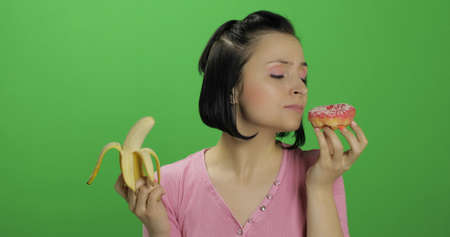 Starting healthy eating. Say no to junk food. Beautiful sad young girl on a chroma key background holds donut in one hand and banana on other. Cute woman choice donut or banana to eatの写真素材
