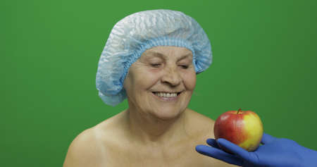 Smiling elderly female lady in protective satisfied with fruit way of treatment. Doctor shows her an apple and patient like this. Concept of healthy eating. Green screen. Chroma keyの写真素材