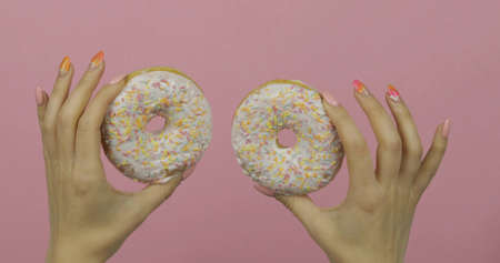 Womens hands holding two white, delicious, tasty and fresh donuts on pink background. Bright and colorful sprinkled sweet donut in girl handの写真素材