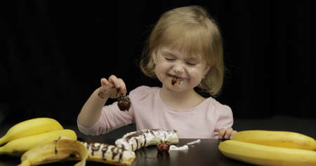 Beautiful, happy child eats strawberry with melted dark chocolate syrup and whipped cream on black background. Teen blonde girl in pink shirt making faces. Cute smiling. Dirty faceの写真素材
