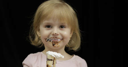 Beautiful, happy child eats banana with melted dark chocolate syrup and whipped cream on black background. Teen blonde girl in pink shirt making faces. Cute smiling. Dirty faceの写真素材