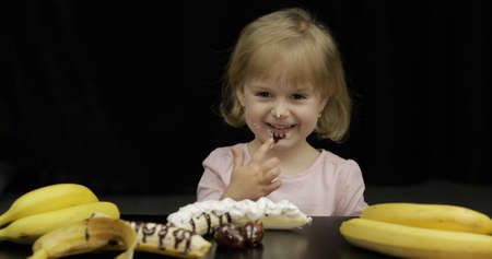Beautiful, happy child eats melted dark chocolate syrup and whipped cream on black background. Teen blonde girl in pink shirt. Cute smiling. Dirty faceの写真素材