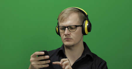 Guy using smart phone in wireless yellow headphones. Portrait of attractive man using mobile phone in headphone while sitting near green screen. Chroma keyの写真素材