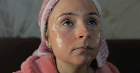 Beautiful cheerful woman sitting on the sofa at home and watching a late night movie at TV, relaxing. In a bathrobe with a towel on her head, the face is covered by facial maskの写真素材