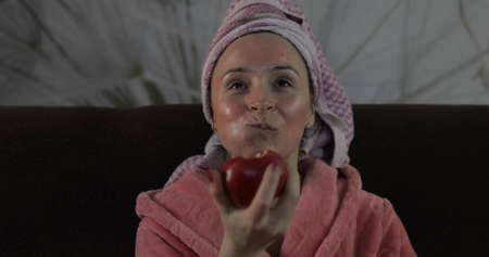 Beautiful cheerful woman sitting on the sofa at home and watching a late night movie at TV, relaxing, eating an apple. In a bathrobe with a towel on her head, the face is covered by facial maskの写真素材