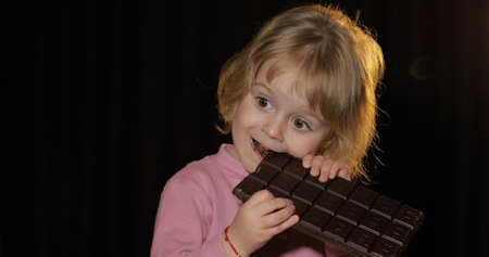 Attractive child eating a huge block of chocolate. Cute blonde girl bite a chocolate. Brown eyes. Pretty little child, 3-4 year old blonde girl. Black backgroundの写真素材