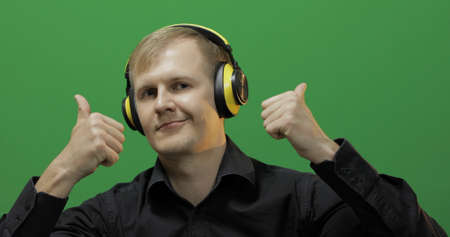 Guy listens to energetic music in wireless yellow headphones and dances. Portrait of attractive man using smart phone for listening the music in headphone while sitting near green screen. Chroma keyの写真素材