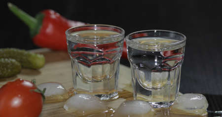 Two shots of vodka in glasses with ice cubes, cherry tomatoes, marinated cucumbers, pepper on the...