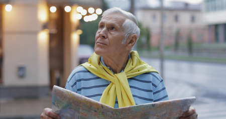 Senior male tourist exploring town with a map in hands and looking for the route while traveling in...