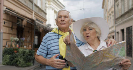 Senior two male and female tourists standing in town with a map in hands and looking for the route...