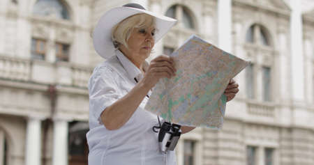 Senior female tourist exploring town with a map in hands and looking for the route while traveling in Lviv, Ukraine. Elegant woman in hat with binoculars. Vacation conceptの写真素材