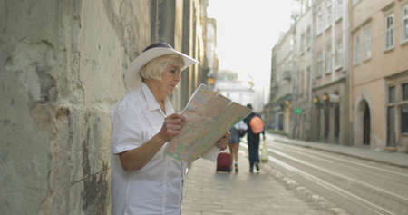 Senior female tourist exploring town with a map in hands and looking for the route while traveling in Lviv, Ukraine. Elegant woman in hat. Vacation conceptの写真素材