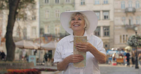 Senior female tourist exploring town with a map in hands and looking for the route while traveling in Lviv, Ukraine. Smiling. Elegant woman in hat. Vacation conceptの写真素材