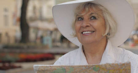 Senior female tourist exploring town with a map in hands and looking for the route while traveling in Lviv, Ukraine. Smiling. Elegant woman in hat. Vacation conceptの写真素材