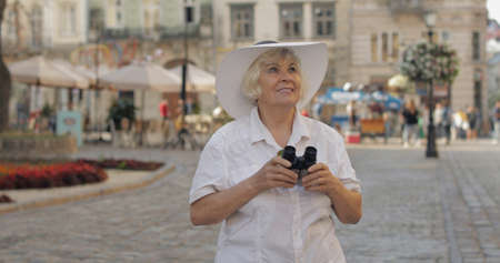 Senior female tourist exploring town and looking in binoculars. Traveling in Lviv, Ukraine. Elegant woman in hat. Vacation conceptの写真素材