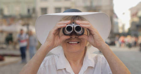 Senior female tourist exploring town and looking in binoculars. Traveling in Lviv, Ukraine. Elegant woman in hat. Vacation conceptの写真素材