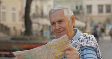 Senior male tourist exploring town center with a map in hands and looking for the route while traveling in Lviv, Ukraine. Smiling. T-shirt and sweater in his neck. Vacation conceptの写真素材