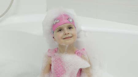 Attractive four years old girl takes a bath with bath foam in pink swimwear and swimming cap. Bath...