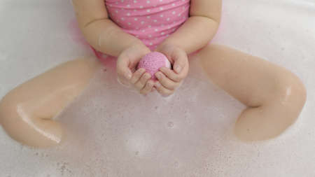 Hands of little girl in pink swimwear puts bath bomb to the water in bathroom. Ball of bath salt...