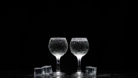 Two frozen vodka shot glasses with ice cubes against black background. Clear transparent alcohol drink rum tequila in shot-glassesの写真素材