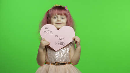 Smiling little girl princess holds a cut pink paper heart with text message about mom in her hands....