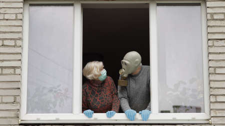 Old grandfather in gas mask and grandmother in protection mask stay at window. Isolated at home on quarantine. Virus outbreak. Elder man, woman. COVID-19 pandemic coronavirus. Social distancingの写真素材