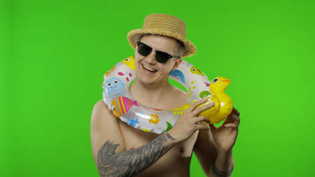 Shirtless young happy man tourist with swimming ring on shoulders play with inflatable duck toy....